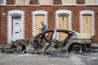 A burnt-out car in front of boarded up houses