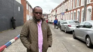 PA Media A man standing in a street lined with terraced houses. He is wearing a brown anorak and pink shirt and wearing dark-lensed glasses. He is balding, and has some hair towards the back of his head. On the right side there is a line of cars parked in front of the houses. On the left the kerbstones are painted red, white and blue. It is an overcast day.