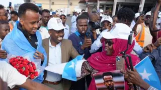 Omar Artan, who has a light blue Somali flag wrapped around his shoulders and is holding a bunch of bright red flowers, is ushered through a packed crowd of people at Mogadishu Airport by a smiling man in a suit who has a white cap on his head bearing a photo of Artan. Various cameras and phones can be seen held up to record proceedings while a lady in a pink headscarf, sunglasses and white Artan cap reaches forward to hand Artan a picture of himself