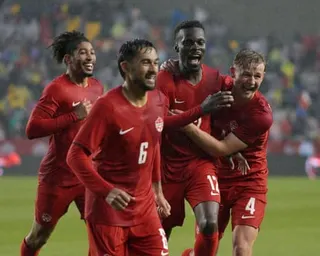 Four Canadian soccer players in red uniforms celebrate together on the field, smiling and embracing