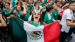 A person wearing green holds a Meican flag to the camera, with a crowd of people celebrating visible behind her.