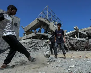 A boy kicks a football towards others next to destroyed buildings