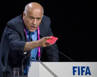 Jibril Rajoub leans forward at a podium holding a red card, speaking at a FIFA event