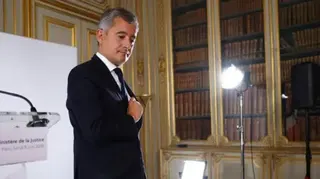 Reuters The minister is at a press conference, where he is wearing a dark suit and tie and is holding a light brown notebook in his left hand. There are some camera lighting stands to his left and the image shows him reaching into his suit pocket with his right hand