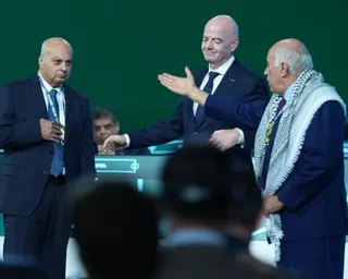 Head of Israel’s football federation, Basim Sheikh Suliman, Fifa boss, Gianni Infantino and Jibril Rajoub in suits stand on a stage at a football congress. Jibril Rajoub, wearing a black-and-white keffiyeh scarf, gestures across the stage.