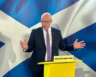 John Swinney speaking in front of a Scotland flag, behind a lectern reading: 'On Scotland's side'