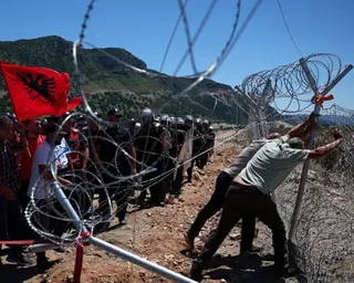 Protesters hold an Albanian flag as two men struggle to destroy a razor-wire fence and police in riot gear stand by.