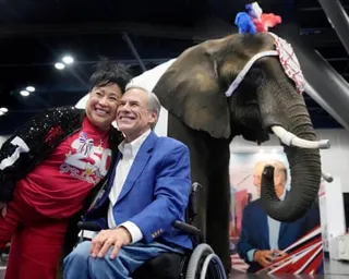 Man in blue blazer in wheelchair with arm around woman in red smile for picture, with African elephant in red, white and blue headdress behind them.