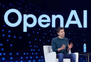 Sam Altman, wearing a long-sleeve blue shirt, sits in a chair in front of a screen that says, OpenAI.