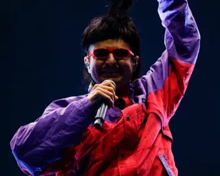 Oliver Tree holds a microphone and raises one arm while wearing a purple and red jacket and red sunglasses