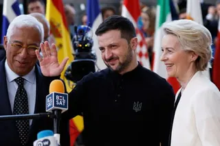 Volodymyr Zelenskyy waves and stands next to Ursula von der Leyen at a media event with flags behind them