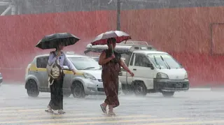 Two people walking across a road in heavy rain in Myanmar, with traffic in the background. They are holding umbrellas, as the rain bounces off the road crossing