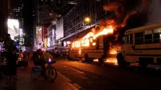A yellow school bus is set alight with fire and smoke protruding from the vehicle at night time in Manhattan, New York City. Bystanders are gathered on the pavement opposite where the bus is parked, some taking videos on their phones. One bystander is sat on a bike.