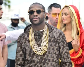 Floyd Mayweather in sunglasses and multiple gold chains wears a patterned shirt with people in the background
