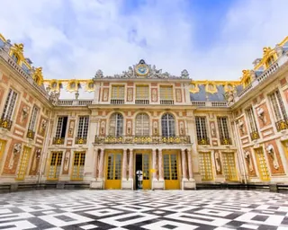 A black-and-white tiled floor leads to three grand doorways in an ornate palace facade beneath a blue sky