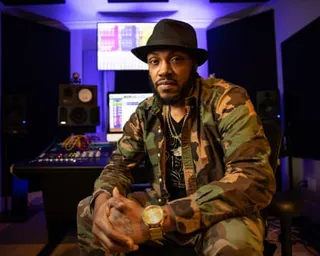 Rapper Mystikal in a camouflage jacket and fedora hat sits in a recording studio with large speakers