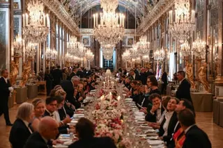 Guests in formalwear sit around a very long table that recedes into the distance beneath rows of chandeliers and surrounded by mirrored walls
