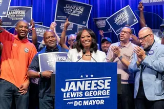 A woman smiles behind a lectern while a crowd of supporters holding signs cheers