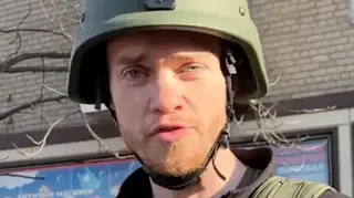 Chris Parry is looking at the camera, you can just see his face and part of his shoulders . He has blue eyes and a short ginger beard, he is wearing a green helmet with a black chin strap and green and black clothing. In the background you can see a blue billboard with Ukrainian writing on it and a tree with bare branches. The sunlight is across Chris' face.