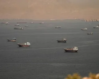 Numerous cargo vessels are scattered across the Strait of Hormuz under a hazy sky