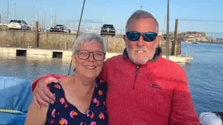 A man wearing a red quarter-zip sweater has his arm around a woman wearing glasses and a navy blue sleeveless top.