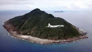 一架白色飛機在空中傾斜飛行，掠過鬱鬱蔥蔥的丘陵島嶼，該島嶼屬於尖閣／釣魚島群島的一部分。