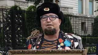 A man wearing an old-style Russian hat with a Soviet badge holds the top of a framed picture and wears a jacket crammed with medals