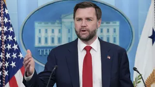 Vice President JD Vance, in a navy suit and red tie stands at a podium, flanked by American and white flags, speaking before a blue backdrop with a circular seal at a press briefing.