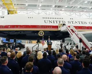 Donald Trump stands at a lectern in front of a large crowd in a large hangar, with a massive white jet behind him.