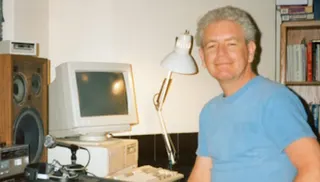 A man with curly gray hair wearing a blue t-shirt smiles at the camera, positioned to the right of a desktop computer setup. The setup includes a beige monitor, a beige computer tower, a white keyboard, a microphone on a stand, headphones, and a large wooden speaker. A desk lamp is positioned between the computer and the man. A bookshelf filled with books is visible in the background.