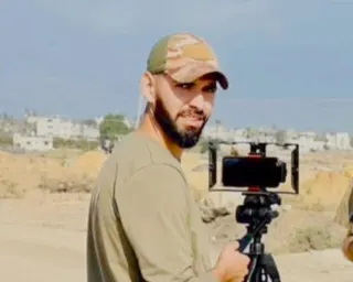 Ahmed Wishah wearing a camouflage cap holds a smartphone on a tripod in a sandy area