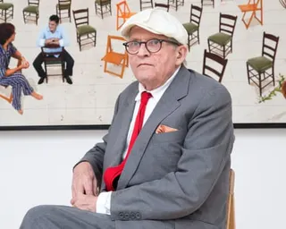Hockney in a grey suit, red tie, and white cap sits in a chair in front of artwork showing chairs