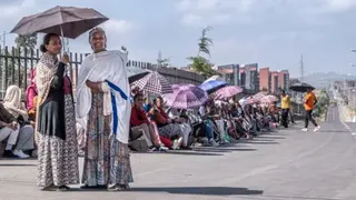 AFP via Getty Images Two women in long dresses and wearing shawls stand under an umbrella - behind them a long line of voters, many under umbrellas, sit on a pavement by a tarmac road in Addis Ababa - 1 June 2026
