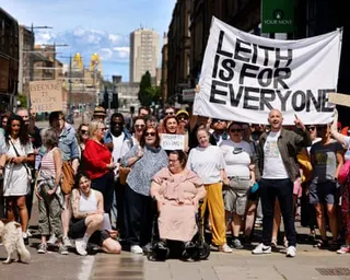 A diverse crowd of protesters poses on a street holding a large banner reading 'LEITH IS FOR EVERYONE'