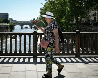A woman in a sun hat uses a hand fan as she crosses a bridge over the Seine in Paris