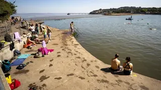 Swimmers sitting beside the marine lake in Clevedon, with various towels left around the outside of the pool, and the pier visible on the horizon in the background.