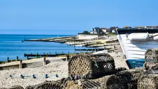 A pile of lobster cages and the bow of a boat resting on a pebble beach. Houses and groynes along the beach.