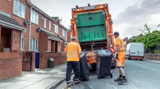 Two refuse collectors in orange high-vis. They are moving two black bins towards the back of a truck on a street lined with houses on the left.