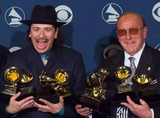 Clive Davis and Santana celebrate their success at the 2000 Grammy Awards, they are both wearing black suits and holding several Grammy trophies each.