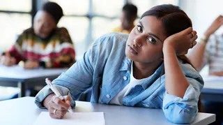 Portrait of young female student wearing a blue shirt leaning on hand while writing exam at desk in classroom
