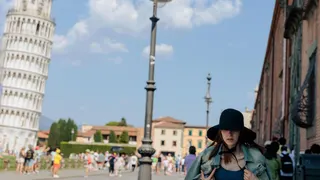 A woman wearing a large sunhat and shielding herself from the sun walks past the leaning tower of Pisa