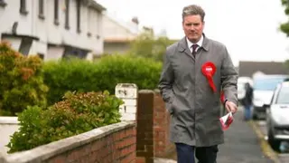 Keir Starmer walking on a residential street towards the camera in 2017. He is wearing a red labour rosette and a rain mac