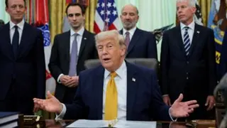 A man in a suit sits at a desk with four men standing behind him, his hand held open.