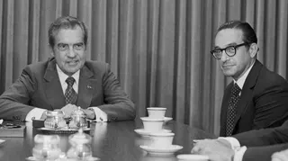 A black and white photo of Richard Nixon and Alan Greenspan. They are sitting at a table which is covered with tea cups.