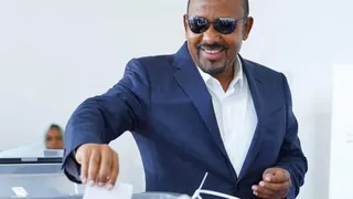 Abiy Ahmed in a navy suit, white shirt and aviator glasses smiles as he places a ballot in a ballot box. Behind him a female electoral official can be seen.