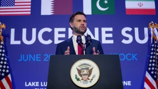 JD Vance speaking at a podium - he is holding his hands to make a gesture while speaking and looking out. He is flanked on either side by American flags, with the flags of Iran, Qatar, Saudi Arabia and the US visible on a graphic behind him.