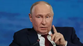 President Putin lifts his finger as if to make a point as he speaks into a microphone. He is dressed in a navy blue suit, white shirt and red patterned tie and is speaking in front of a marine blue backdrop