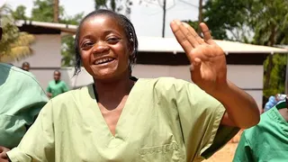 A health workers wearing green scrubs smiles and dances as she waves hr left hand in the air.