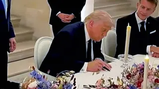 Donald Trump signing a document  at a table with white table cloth and flowers. Macron is seen to his right watching