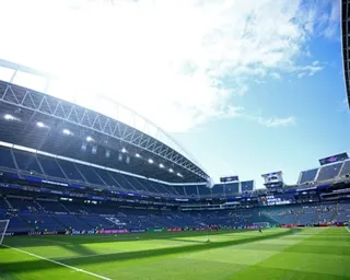 An empty football stadium with blue seats, a covered stand, and bright green pitch under a blue sky